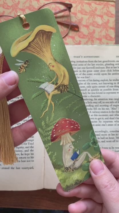 Video: Mushroom Bookclub bookmark held over an open book, showing two reading mushrooms and a bee on green