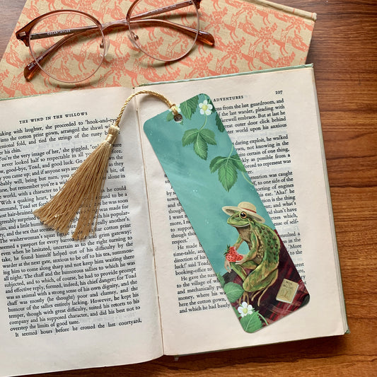Froggie Picnic bookmark by Anna Seed on an open book by glasses, a frog in a sunhat eating a strawberry on a rug
