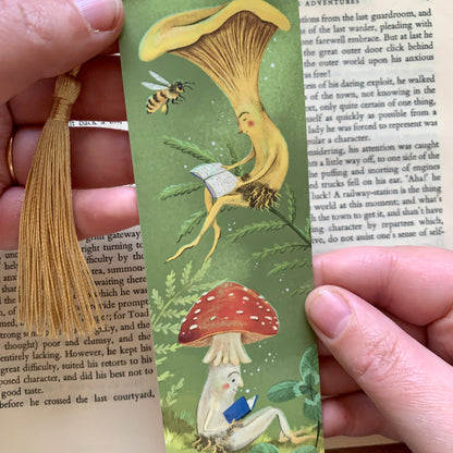 Close-up of the Mushroom Bookclub bookmark held in hand, a yellow chanterelle and spotted toadstool reading books