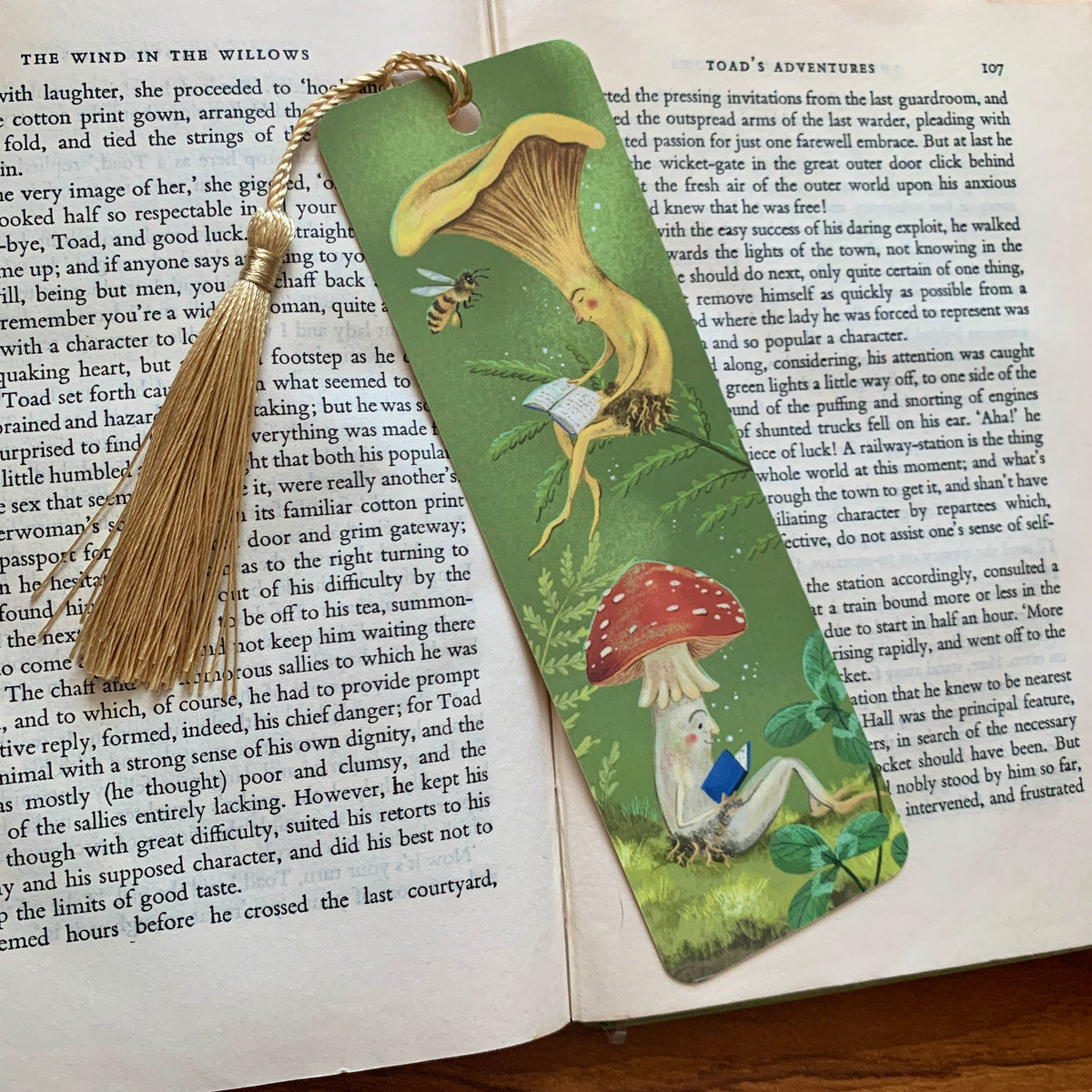 Mushroom Bookclub bookmark with a gold tassel lying on an open book, two mushrooms reading among ferns