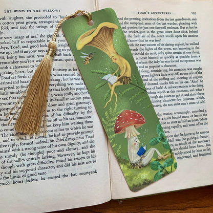 Mushroom Bookclub bookmark with a gold tassel lying on an open book, two mushrooms reading among ferns