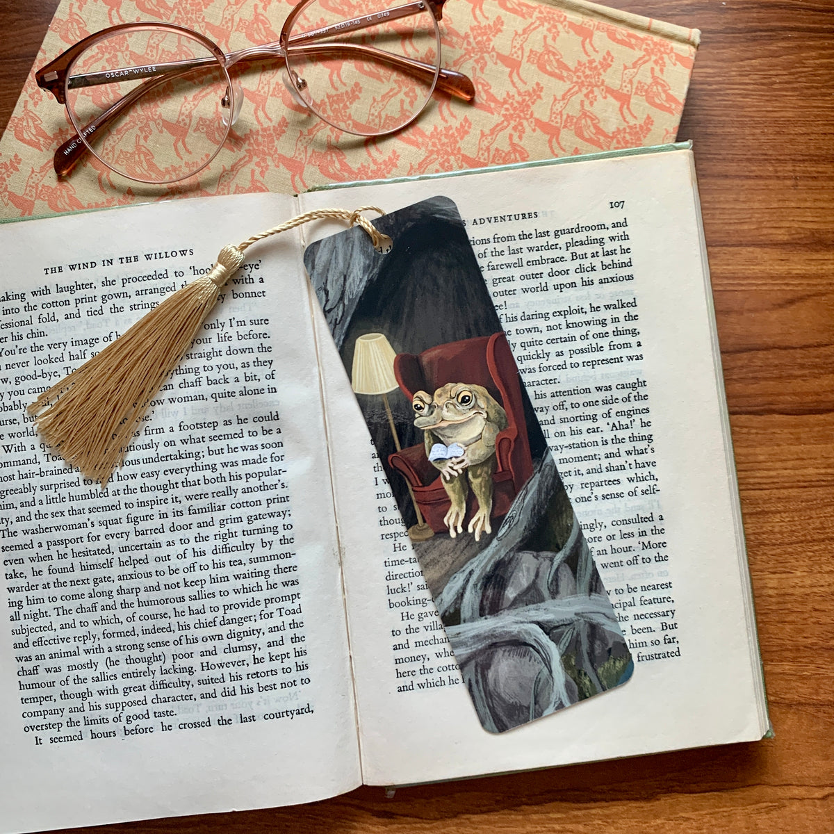 Reading Toad bookmark by Anna Seed on an open book beside glasses, a toad reading in a red armchair by lamplight