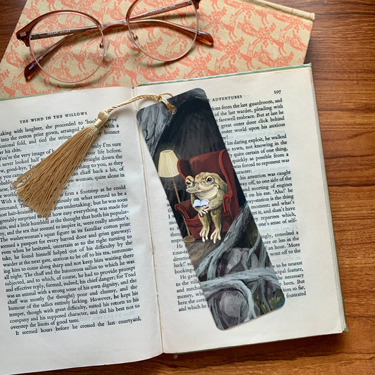 Reading Toad bookmark by Anna Seed on an open book beside glasses, a toad reading in a red armchair by lamplight