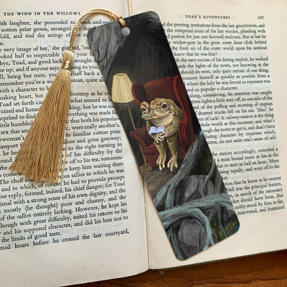 Reading Toad bookmark with a gold tassel lying on the pages of an open book, the toad reading among grey roots