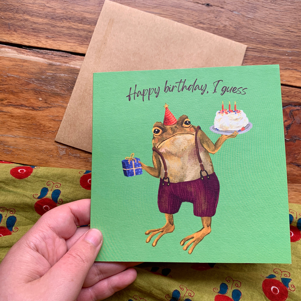 Happy birthday, I guess card by Anna Seed: a grumpy toad in a party hat holding cake and a gift, held in hand