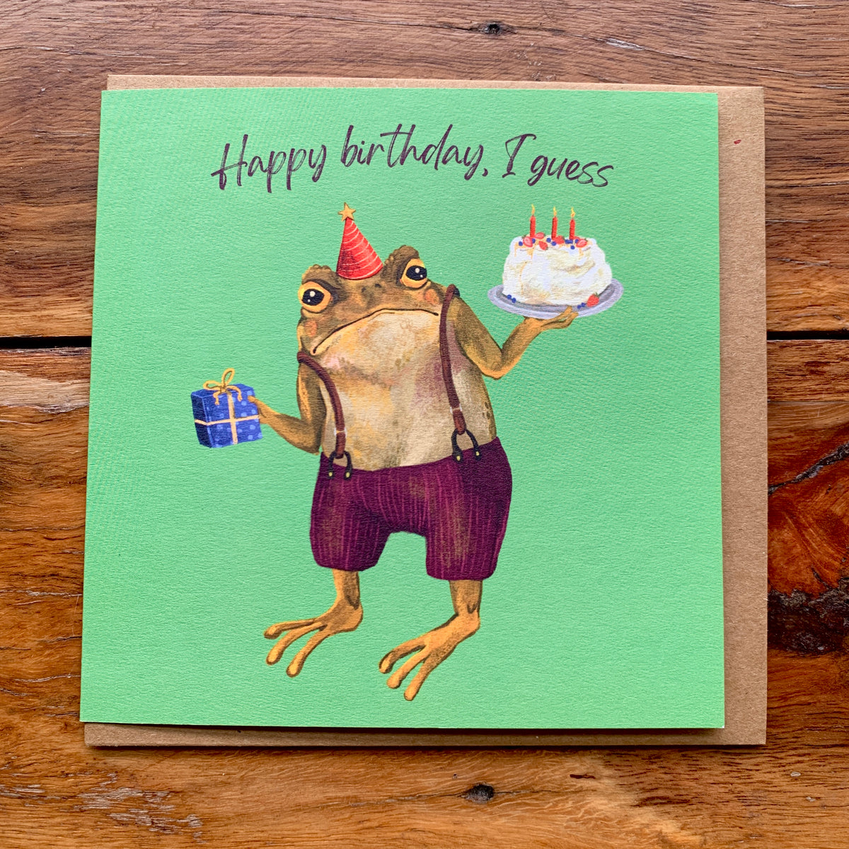 Happy birthday, I guess card with an unimpressed toad holding a birthday cake on green, flat on a wooden table