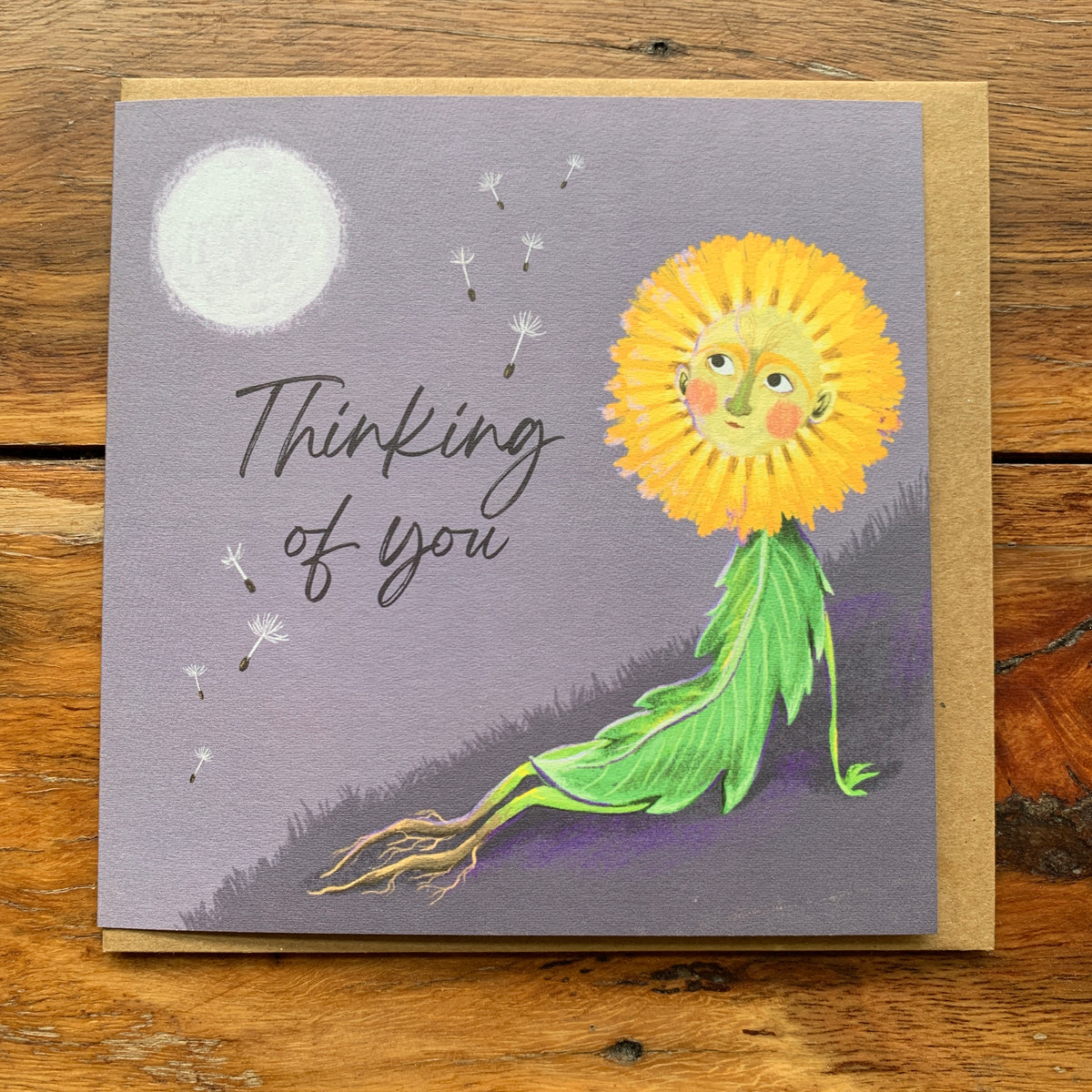 Thinking of you card with a dandelion figure gazing at the moon and drifting seeds on lilac grey, on a wooden table