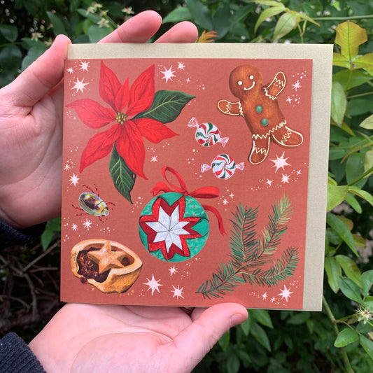 Aussie Christmas red card by Anna Seed: poinsettia, gingerbread man, bauble and mince pie on rust red, held in hands