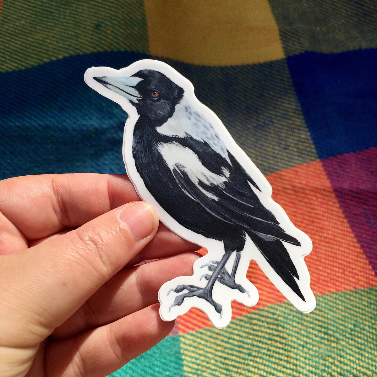 Magpie sticker held in a hand over a colourful blue, orange and green tartan blanket in sunlight
